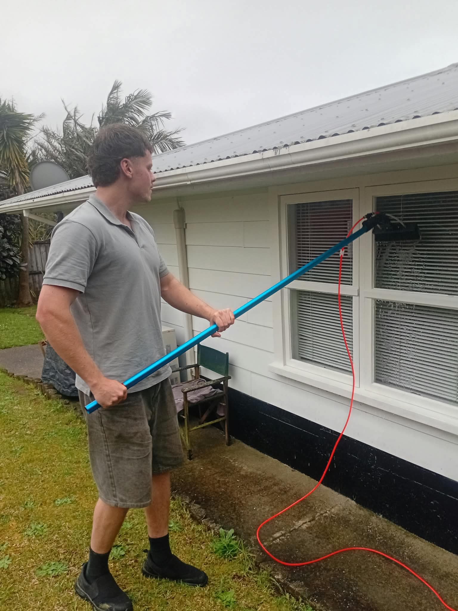 Man cleaning a white house window using a long blue water-fed pole and brush.