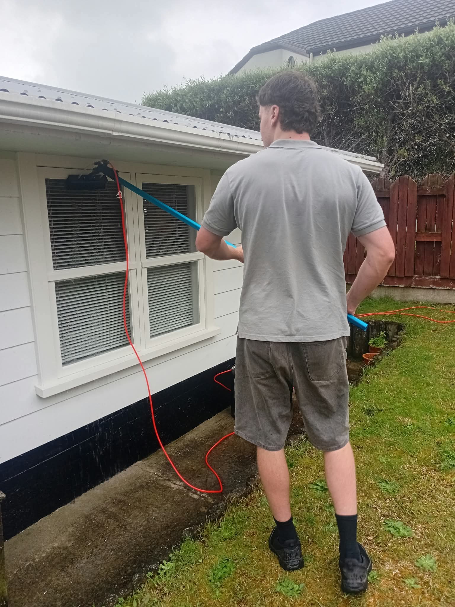 Man cleaning white house windows with a long blue water-fed pole and brush system.